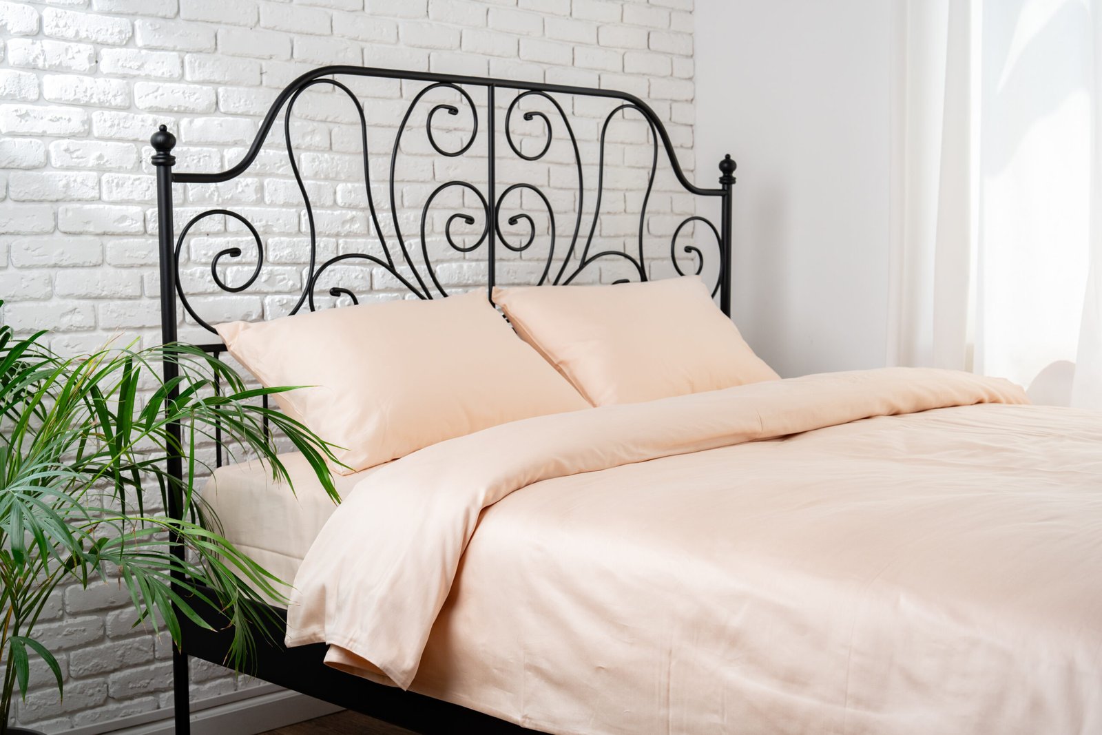 A black metal bed frame with a scrolled headboard sits in a bedroom with a white brick wall and a window letting in natural light.