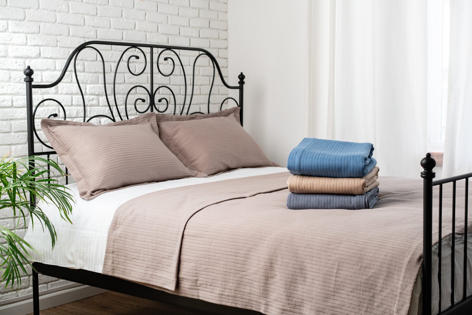 A stack of three blankets, one blue and two beige, sits neatly on a made bed. The blankets are ribbed and folded neatly.